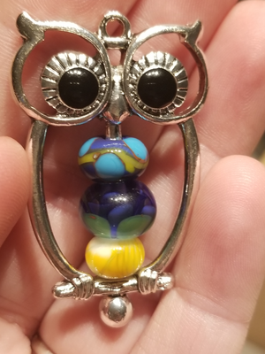 a photo of three handmade glass beads threaded on the central pin of a purchased owl-shaped bead frame.