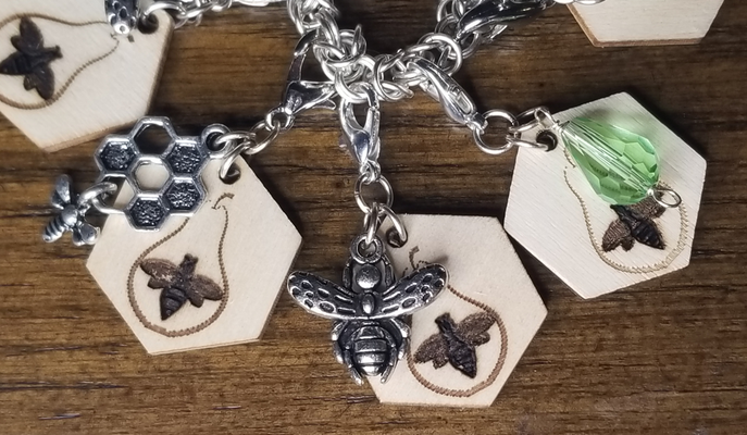 a photo of four composite clip-on charms. Each charm has a hexagonal wooden piece laser-engraved with a bee within 
		a pear outline, plus one of either a silvertone honeycomb charm, a silvertone honeybee charm, or a green teardrop briolette bead.