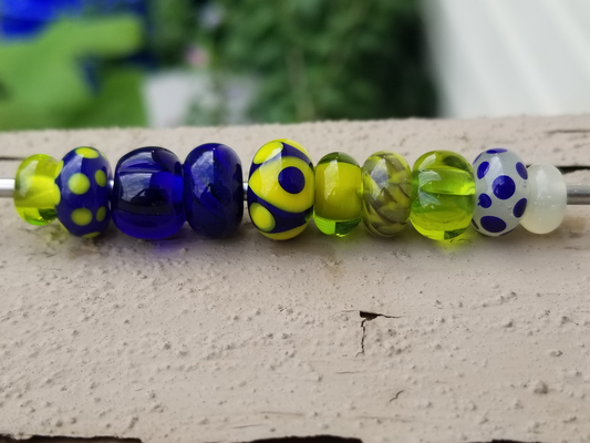 a photo of ten handmade glass beads threaded on a mandrel. Each bead is different, but they all generally 
		share a color theme of blue and yellow