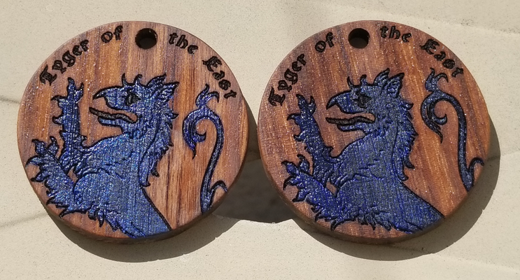 a photo of two handmade wooden medallions, laser engraved with the torso of a rampant tyger plus the words 
		'Tyger of the East'. The tyger is painted blue. The medallions are made of walnut wood.