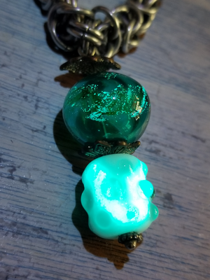 two lampwork glass beads strung together on a headpin, attached to a chain at the top. The upper bead is 
		hollow dark green glass and has swirls of glow pigment across its surface. The lower bead is a white and pale green barrel-shaped bead with raised 
		dots. The surface of the lower bead also has glow pigment, which makes it glow overall when exposed to sunlight or UV source