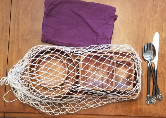 a top-down photo of dishes and associated equipment. A folded purple cloth napkin at the top.
		At the center bottom, a bag of white linen fish-netting encloses an assortement of round and square wooden dishes. To the right 
		of the dishes are a spoon, fork, and knife jumbled together.
