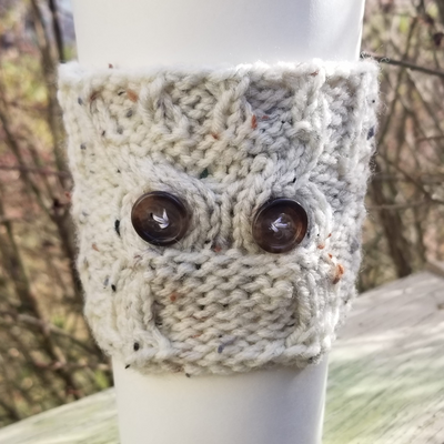 a phogo of a hand-knitted cup sleeve on a typical 20 oz to-go cup from a coffee shop. The sleeve 
		has a cabled pattern and two sewn-on buttons that resemble an owl. The yarn is speckled oatmeal in color.